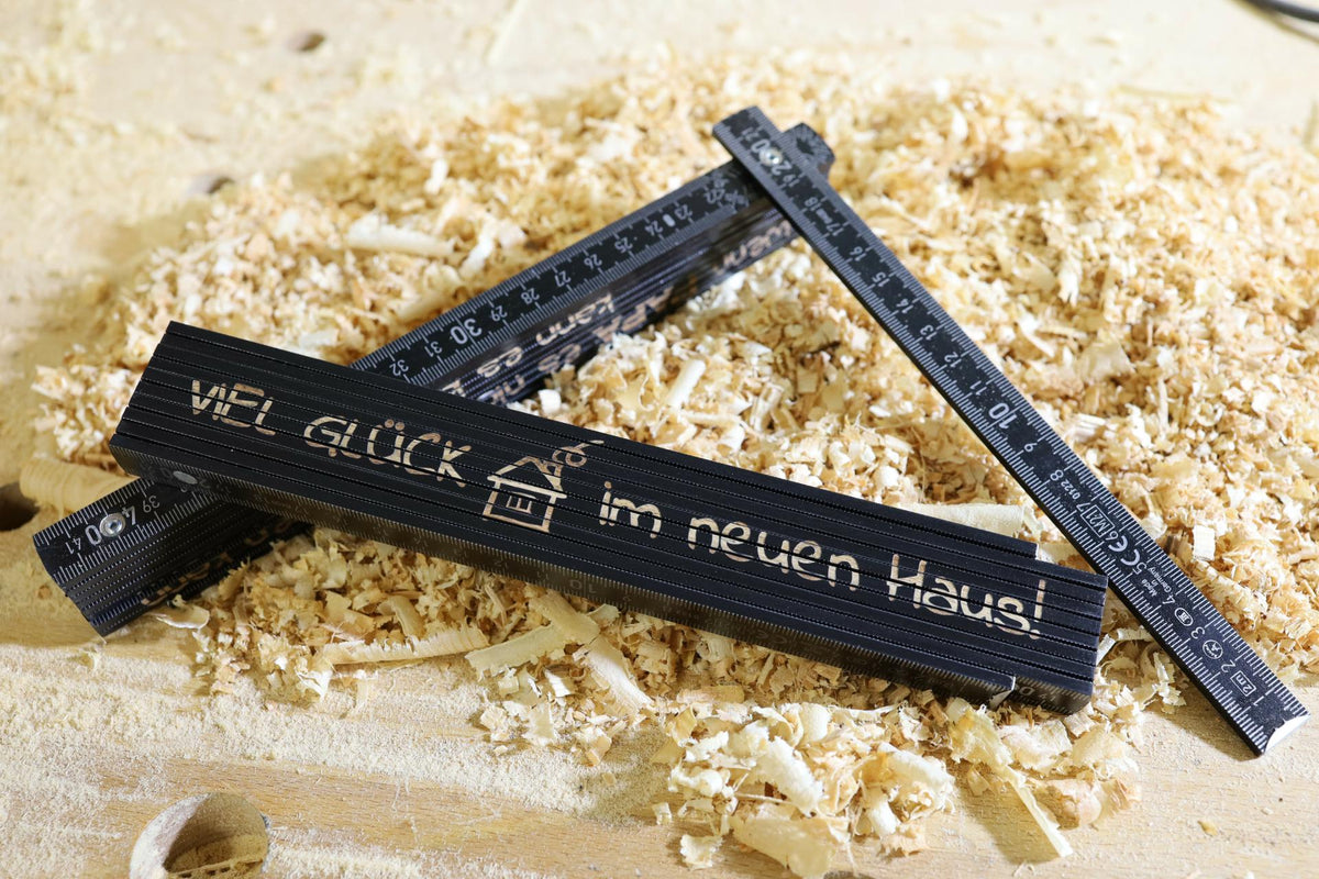 Schwarzer Zollstock aus Holz mit Gravur „Viel Glück im neuen Haus“ auf Holzspänen