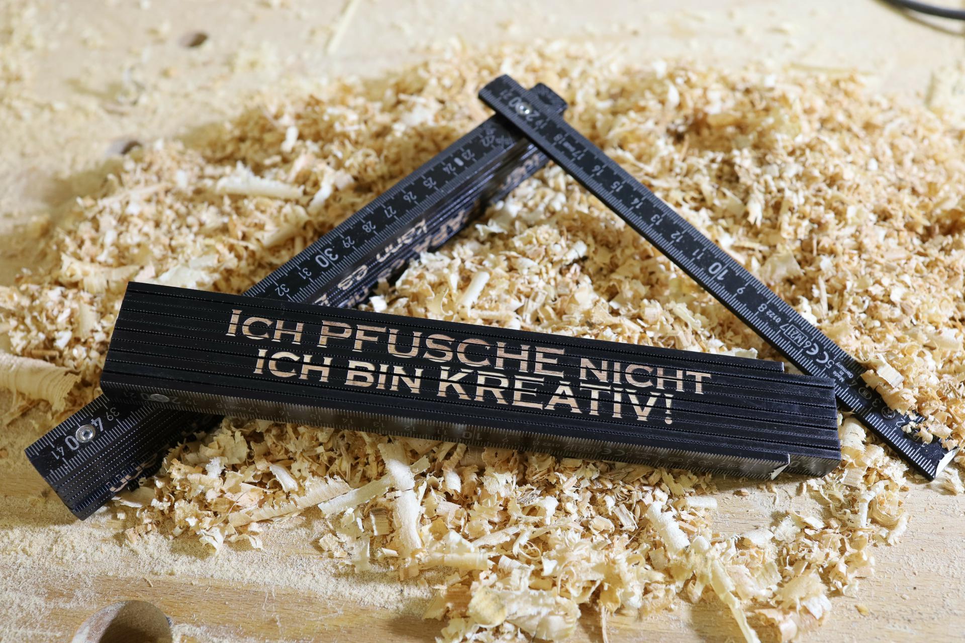 Schwarzer Zollstock aus Holz mit Gravur „Ich pfusche nicht, ich bin kreativ!“ auf Holzspänen
