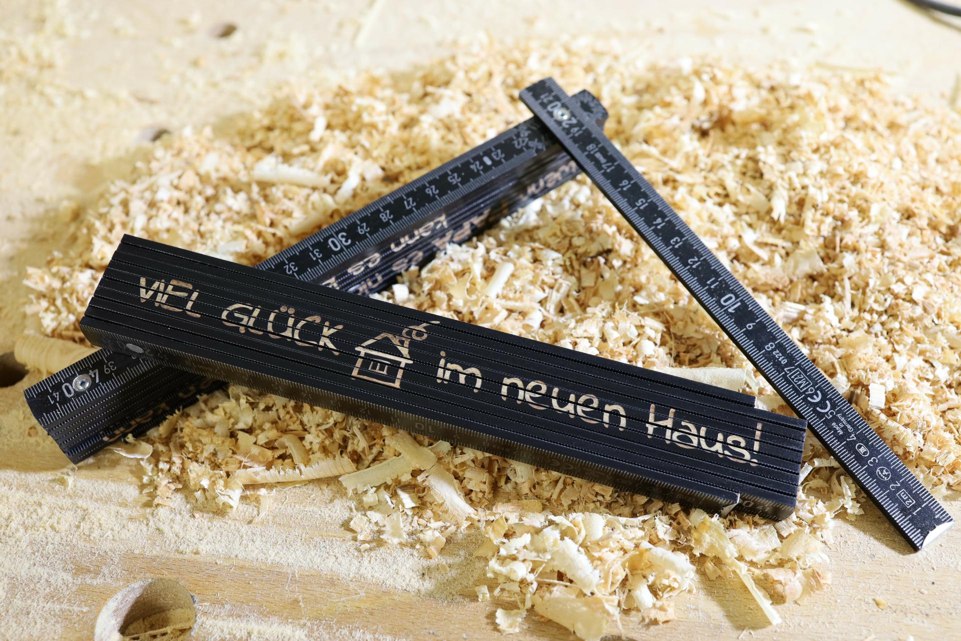 Schwarzer Zollstock aus Holz mit Gravur „Viel Glück im neuen Haus“ auf Holzspänen
